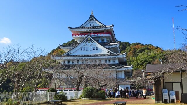 快晴の下にそびえる熱海城と芝生広場、観光客が並ぶ様子